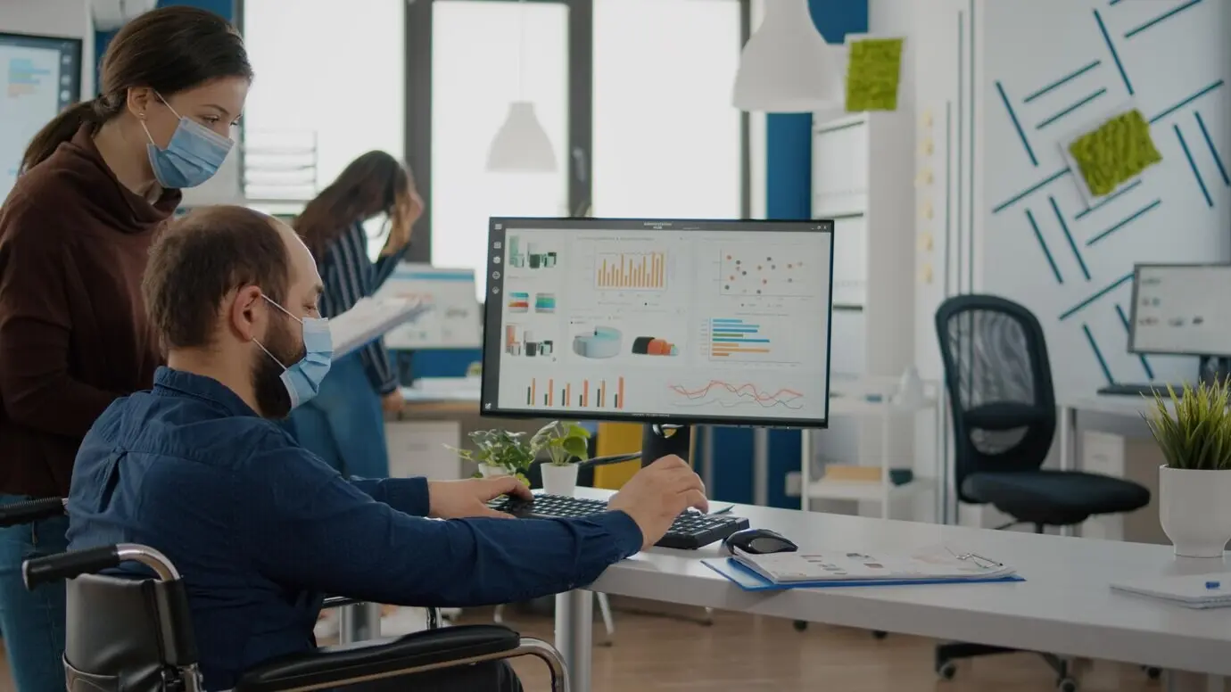 Сотрудник на инвалидной коляске разговаривает с коллегой в офисе стартапа, в масках во время пандемии COVID‑19. Команда финансовой компании в условиях новой нормальности печатает на ПК и проверяет отчеты.